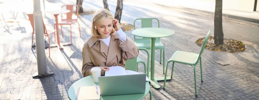 Portrait of young blond woman with laptop, sitting on street, drinking coffee in outdoor cafe, working, wearing wireless headphones, doing homework, freelancing.
