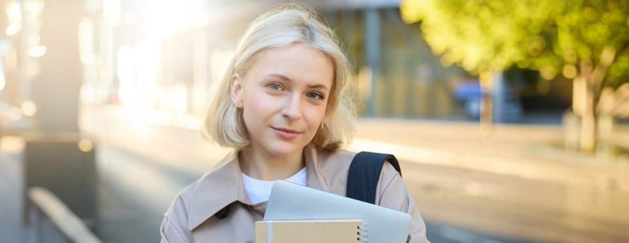Portrait of smiling, carefree young woman in trench coat, holding backpack and notebook, going to college or university, carries study materials for her language courses.