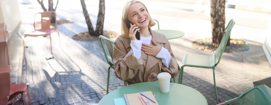 Lifestyle and people concept. Happy beautiful young woman, talking on mobile phone, sitting outside in cafe with cup of coffee, smiling and laughing, chatting about something funny.