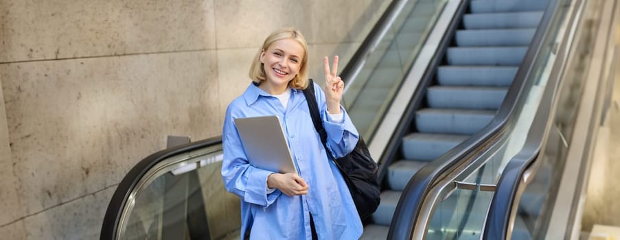 Lifestyle portrait of positive young woman, female student with laptop and backpack, showing peace sign, standing near escalator.