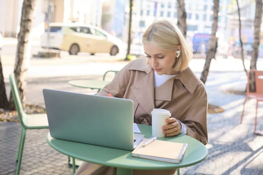 Portrait of young blond woman with laptop, sitting on street, drinking coffee in outdoor cafe, working, wearing wireless headphones, doing homework, freelancing.