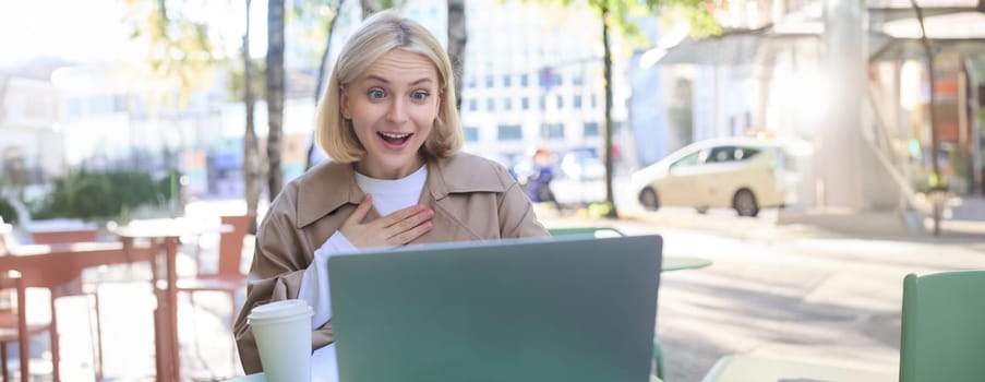 Lifestyle portrait of young surprised woman, looking at laptop with amazed face, sitting in coffee shop with excited face expression, spending time outdoors on sunny day.