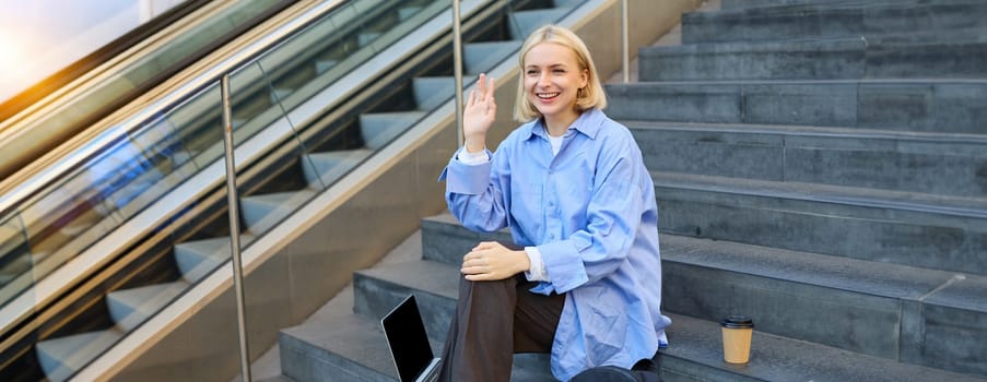 Friendly smiling woman, student waving hand to say hi to friend or classmate, sitting outdoors on campus stairs, resting after working on laptop, drinking coffee.