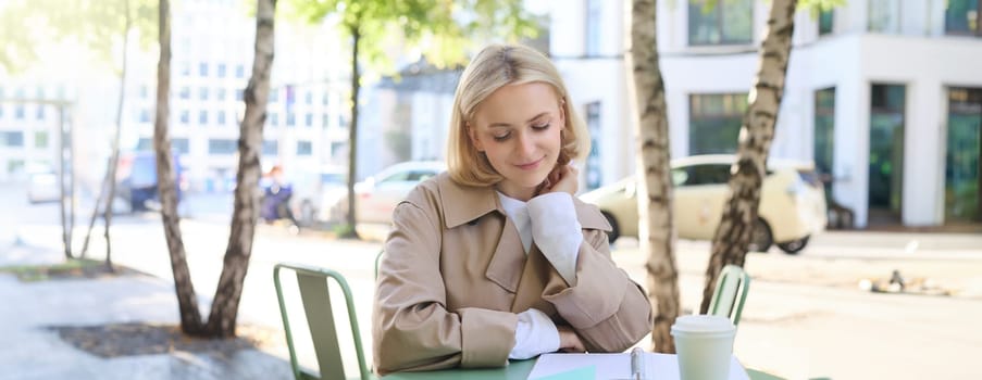 Image of female student, cute girl sitting in cafe, drinking coffee, reading notes in notebook, revising for university lesson, doing homework.