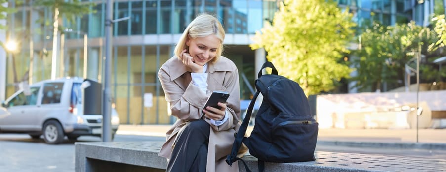 Lifestyle portrait of cute blonde girl with smartphone, sitting on bench with backpack, using mobile phone, reading message.