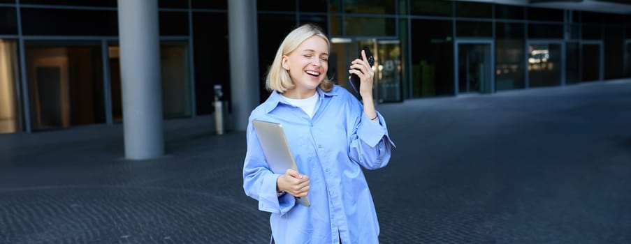 Portrait of carefree, happy young female student, dancing, holding smartphone and laptop, singing with eyes closed and pleased face.