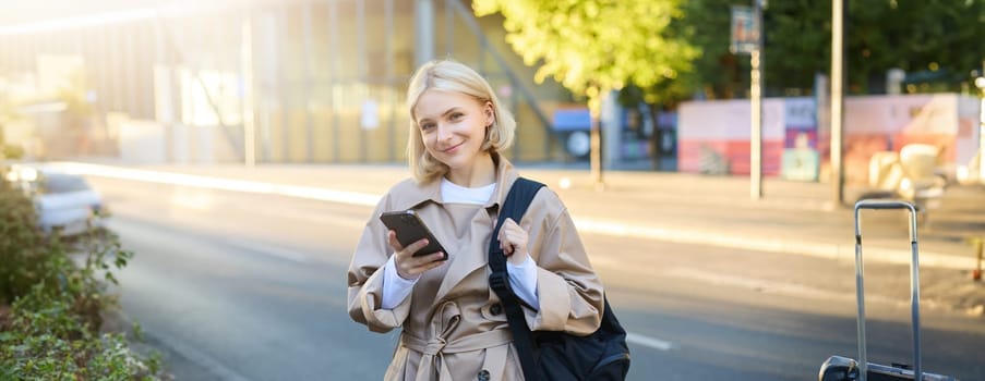 Smiling, beautiful young woman with backpack, holding smartphone, standing on street, using mobile phone app.