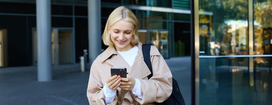 Portrait of young stylish woman walking in city centre with smartphone, using mobile phone app outdoors, waiting for someone, going to work.