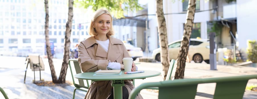 Image of young woman, makes sketches outdoors on street in notebook, drinks coffee and smiles at camera. Lifestyle and people concept