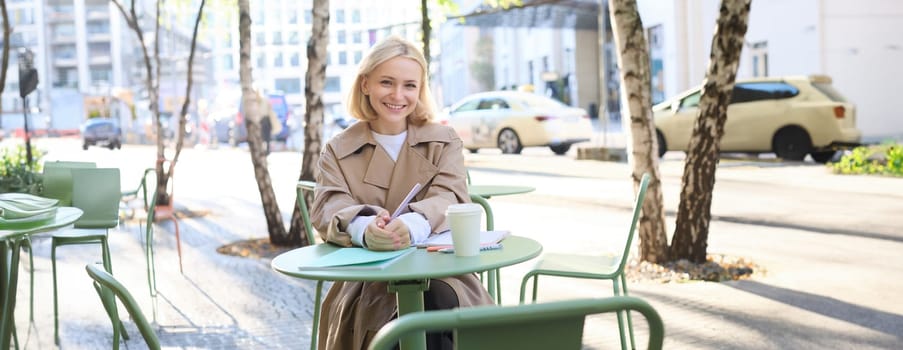 Portrait of young concentrated woman, doing homework in a cafe, writing in notebook, working with documents and drinking coffee outdoors.