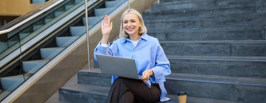 Happy, friendly young woman sitting with laptop on street stairs, waving hand to say hello, working remotely, elearning, connecting to public wifi.