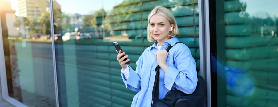 Lifestyle portrait of smiling young female model, student with backpack, waiting for someone on street, standing outdoors with smartphone.