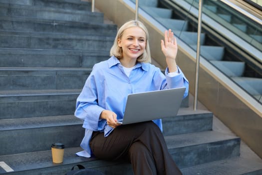 Happy, friendly young woman sitting with laptop on street stairs, waving hand to say hello, working remotely, elearning, connecting to public wifi.