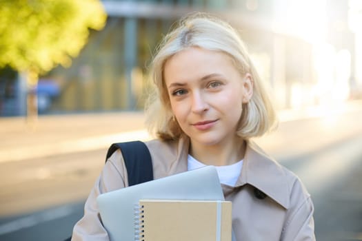 Portrait of smiling, carefree young woman in trench coat, holding backpack and notebook, going to college or university, carries study materials for her language courses.