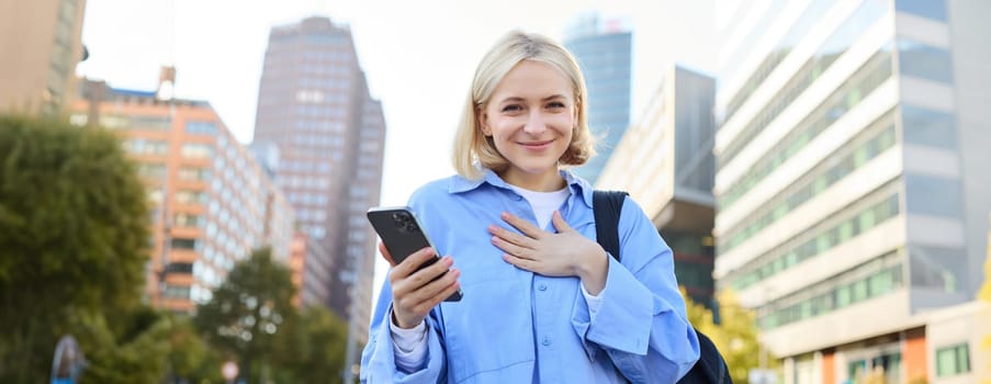 Image of young professional, office manager woman with backpack and smartphone, posing on streets of busy city, holding hand on chest, smiling and looking excited at camera.