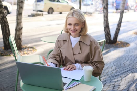 Happy, smiling young woman in trench, sitting in street cafe outdoors, drinking coffee, freelancing, looking at laptop and making notes, writing in notebook, working remotely.