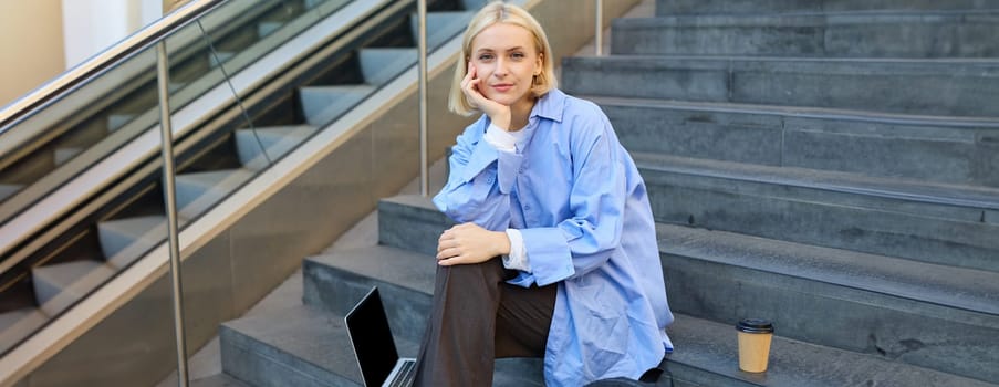 Portrait of young modern woman, student or freelancer, sitting on outdoor stairs, resting in city, has laptop and coffee, looking at camera with confident, smiling face.