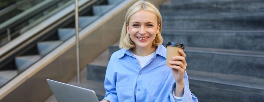 Close up portrait of beautiful blonde girl student, sitting on stairs outdoors, drinking cup of takeaway coffee, working on laptop, studying on street.