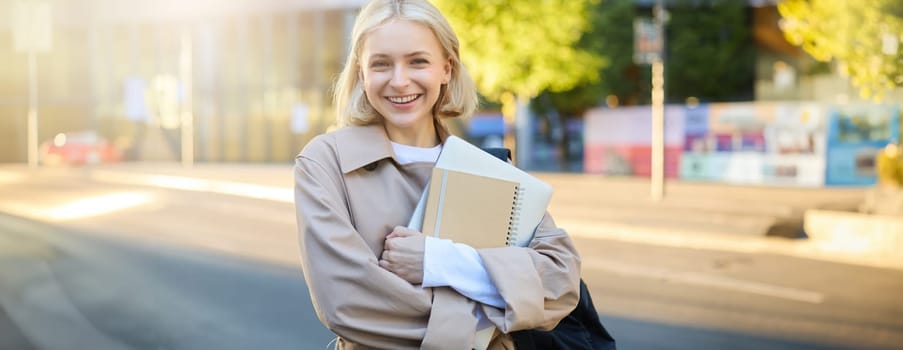 Portrait of beautiful blond girl, college student carries notebooks, holds backpack, smiles and looks happy at camera. University lifestyle concept
