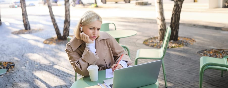 Portrait of blond girl with upset face, listening lecture, attends online course in wireless headphones, sits in outdoor cafe and frowns while makes notes, studying remotely, using laptop.
