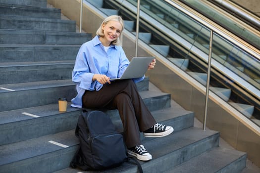 Image of young woman, student studying online, working remotely, sitting on stairs with laptop and drinking coffee in takeaway cup, elearning.