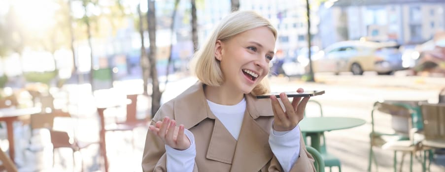 Close up portrait of young smiling woman, lively recording voice message, talking into smartphone microphone, sitting casually in outdoor cafe, gesturing and looking happy.