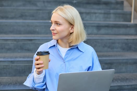 Young woman sitting with cup of coffee and laptop on stairs outdoors, working remotely, freelancing, connecting to public wifi in city to study online.