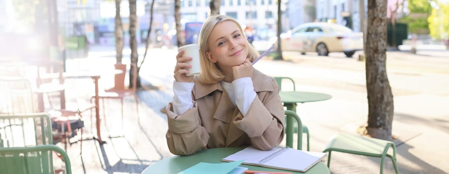 Image of beautiful young blond woman, girl drinking coffee in outdoor cafe, working on university project, doing homework, enjoying sunny weather, smiling at camera.