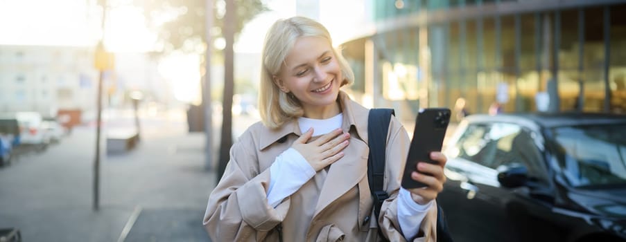 Image of happy, beautiful young woman on street, answers video call, waves hand at smartphone camera, says hello, talks to friend over the mobile app, walking along road.