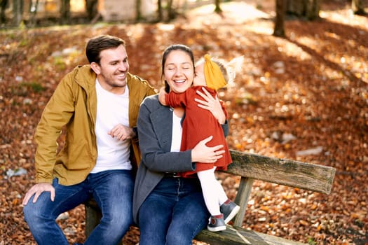 Little girl hugs her mother while standing next to her on a bench next to a sitting dad. High quality photo