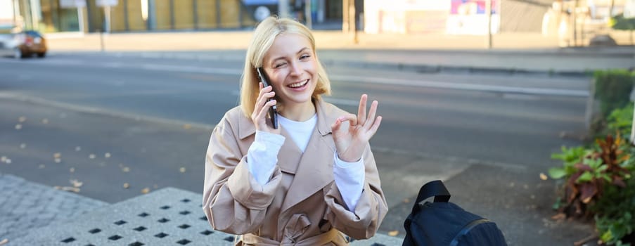 Image of young woman talking on mobile phone, sitting on bench outside, showing okay, ok approval gesture, made a deal over the telephone.