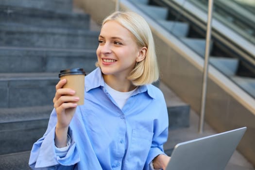 Portrait of blond smiling woman, drinking coffee, using laptop, sitting on campus stairs outside of building, studying, e-learning, connecting to wifi.
