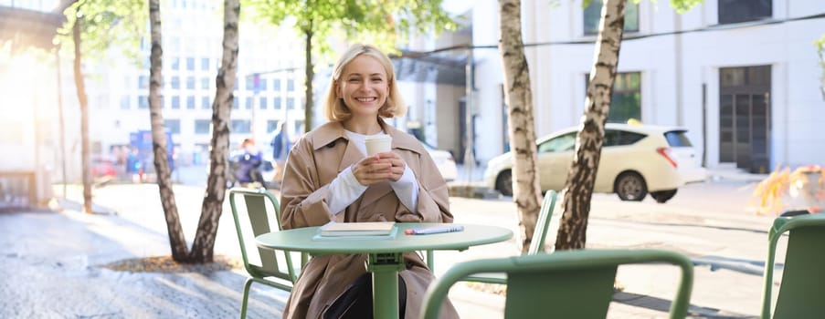 Portrait of stylish modern woman, sitting in an outdoor cafe, smiling and drinking coffee from takeaway cup, wearing trench coat.