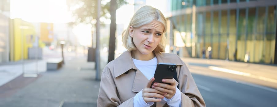 Portrait of young blond woman standing on street, has unsure, doubtful face expression, using mobile phone, showing reluctance.