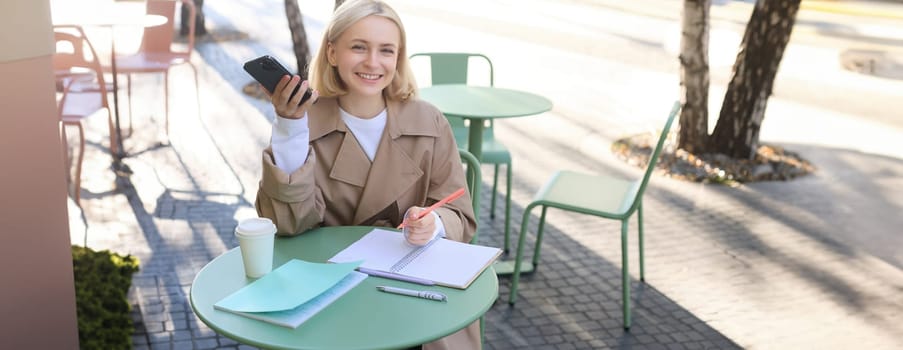 Image of young beautiful girl, student studying, doing homework in cafe, using mobile phone, holding smartphone, smiling.