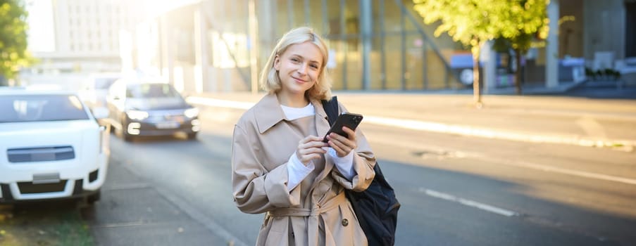 Lifestyle portrait of young woman on street, standing near road with cars, carries backpack and holds mobile phone, requests a ride on smartphone app, smiling and looking happy at camera.