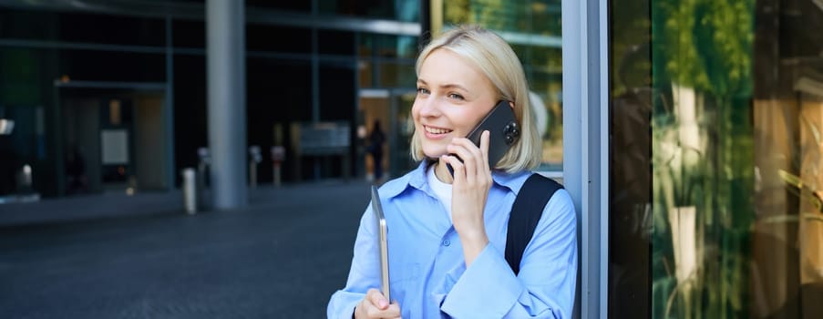 Portrait of young modern woman, office manager near building, standing outside with backpack, laptop, talking on mobile phone, chatting on smartphone. Lifestyle and communication concept