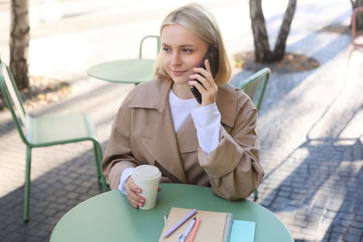 Image of stylish blond woman talking on mobile phone, drinking coffee in cafe outdoors, enjoying warm weather in city centre, answer telephone call on smartphone.