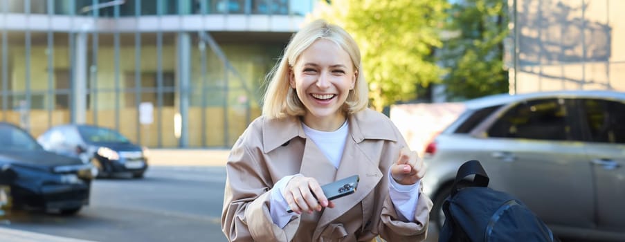Upbeat young woman celebrating, winning and triumphing, sitting on street bench and laughing, holding mobile phone.