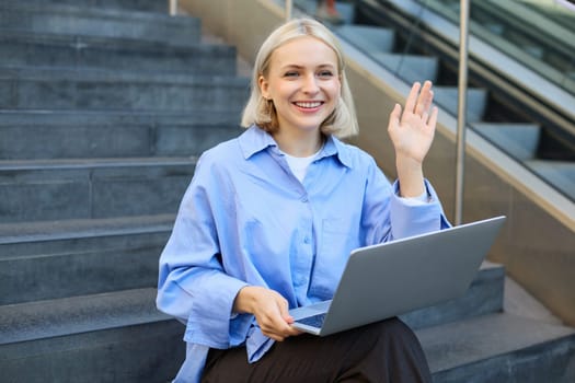 Image of smiling young woman, sitting on stairs outside university or college, waving hand, saying hello to passer-by, working on laptop.