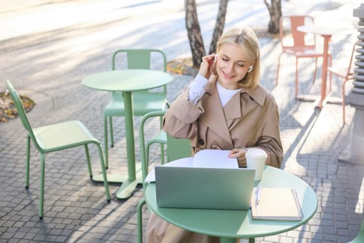 Lifestyle and technology concept. Young blond working woman in trench coat, studying, working on project on laptop, sitting in coffee shop on sunny day, making notes, attending online course.