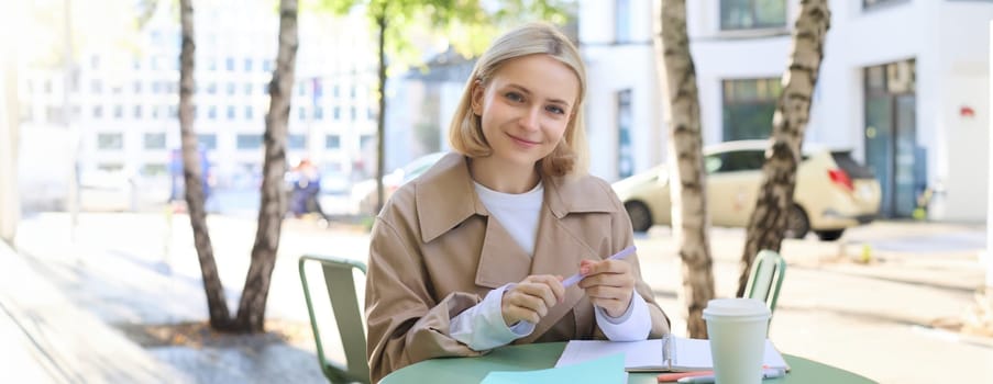 Portrait of carefree, blond cute woman working with documents, drinking takeaway coffee and sitting in cafe outdoors with documents, writing essay, student doing homework.