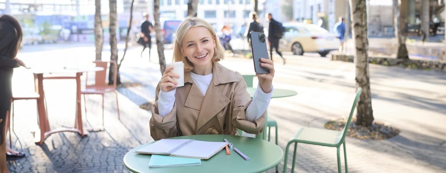 Lifestyle shot of stylish blond girl with smartphone, drinking coffee, posing with her chai for social media, creating content on phone, taking selfie.