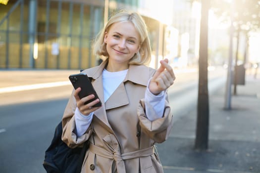 Lovely young female model in trench coat, showing heart gesture with fingers, holding smartphone and smiling at camera, standing on empty sunny street.