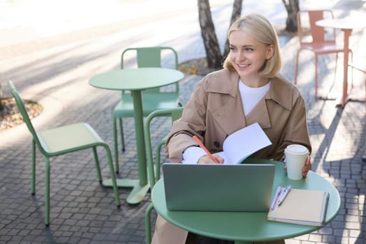Portrait of young smiling, beautiful woman, studying outdoors in cafe, has laptop and coffee on table, writing in journal, doing homework on sunny day.