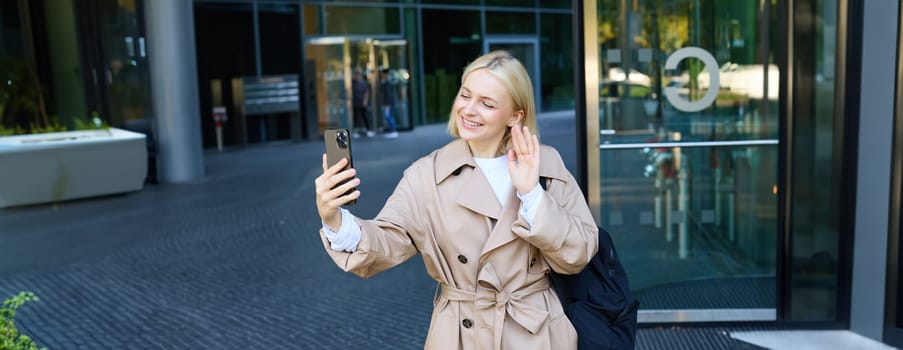 Lifestyle portrait of young modern woman, cute girl video chatting on street, waving hand at her smartphone and smiling, walking on street and talking to friend on mobile phone app.