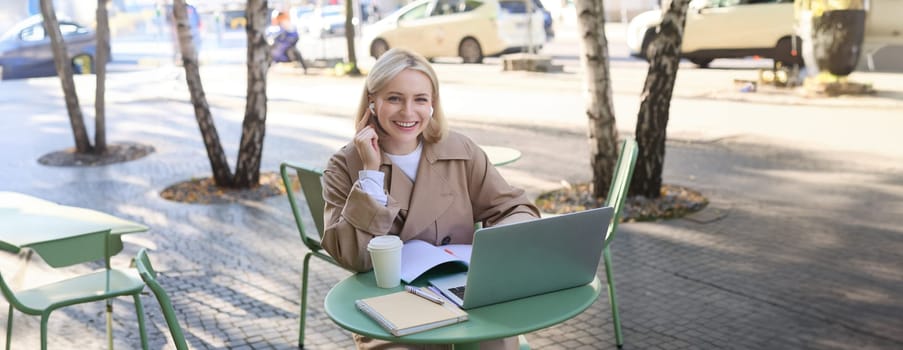 Portrait of beautiful blond woman, wearing wireless headphones, using laptop, studying in outdoor coffee shop, making notes, working on project.