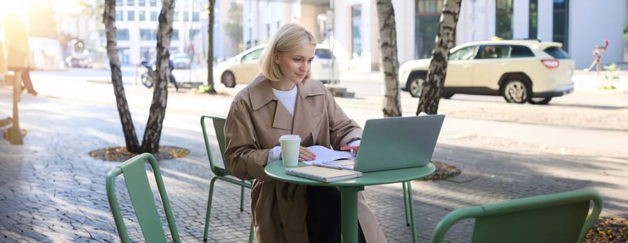 Lifestyle portrait of young modern woman, working on laptop, sitting in quite outdoor cafe, working on project, studying remotely, freelancing while having her coffee, on bright sunny day.