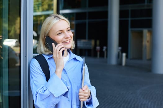 Close up portrait of modern young woman, student standing near building in city centre, talking on mobile phone, having chat on smartphone and smiling.