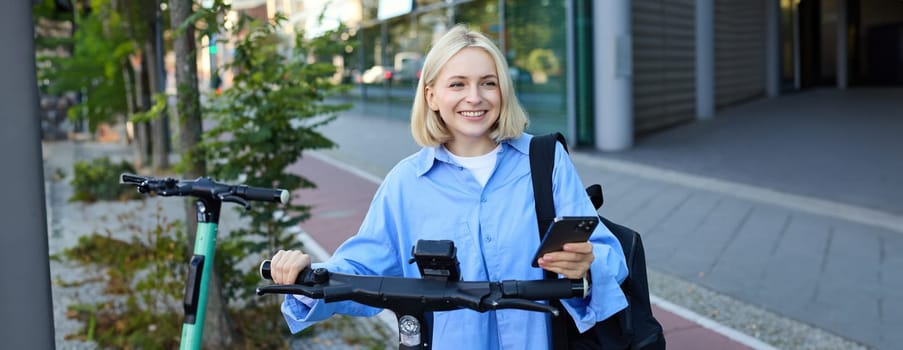 Portrait of young smiling woman, unlocks electric scooter, scans qr code with smartphone app to unlock her ride, standing on street near building.